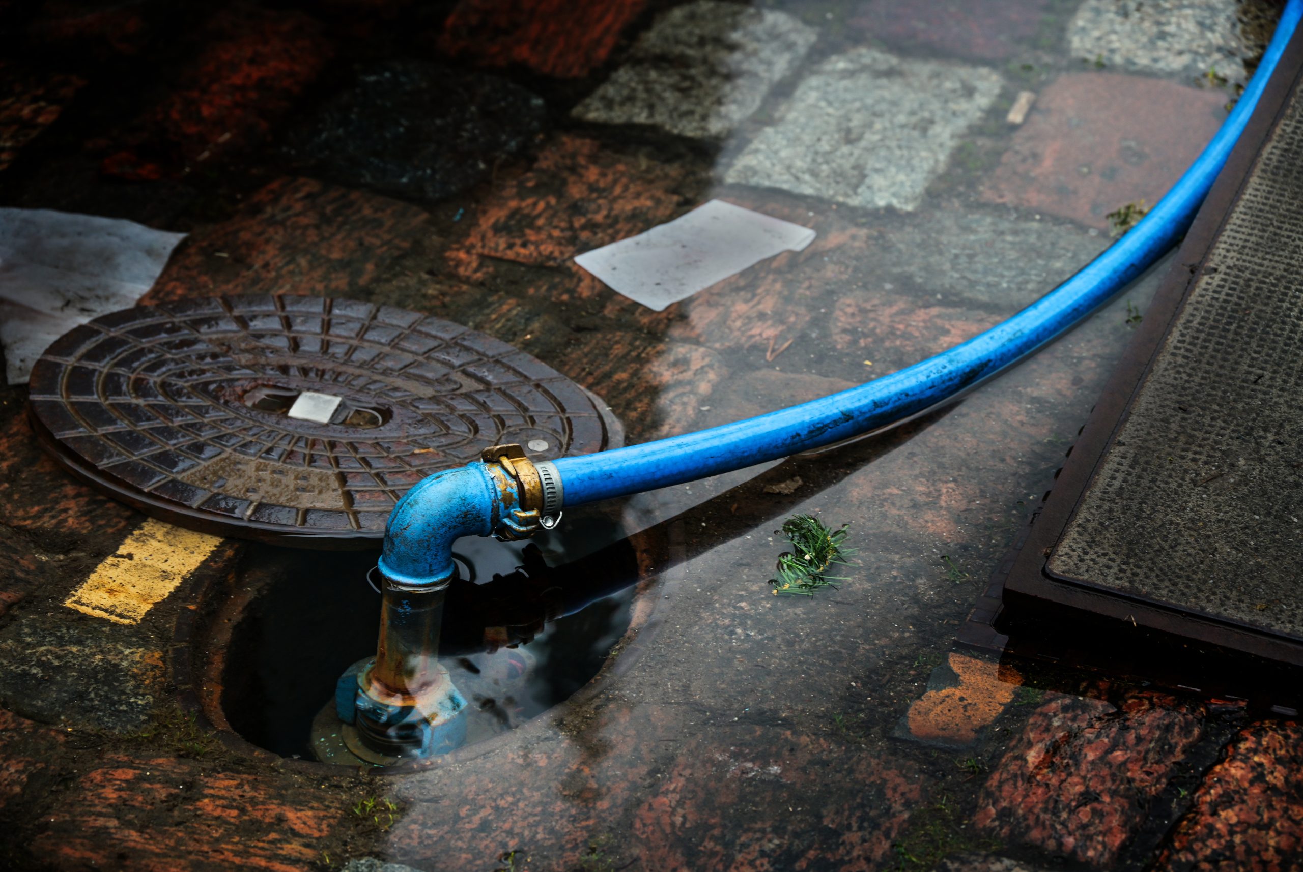 blue water hose connected to a flooded underground hydrant in a cobblestone street, copy space, selected focus, narrow depth of field